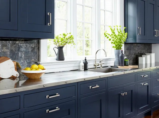 Navy upper cupboards and drawers frame a sink beneath bright windows with pale counters, silver-colored handles and a darker textured backsplash