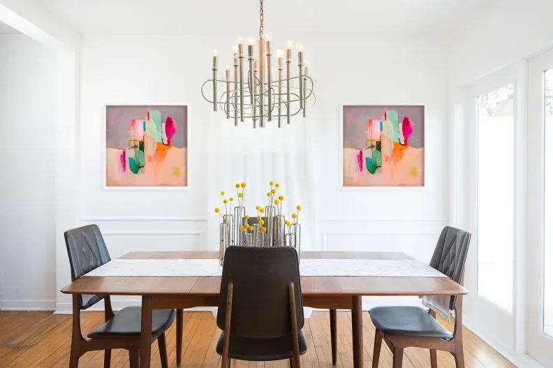 Two colorful abstract pictures in pale frames above a wooden dining table, with black chairs, yellow flowers, and a branching chandelier.