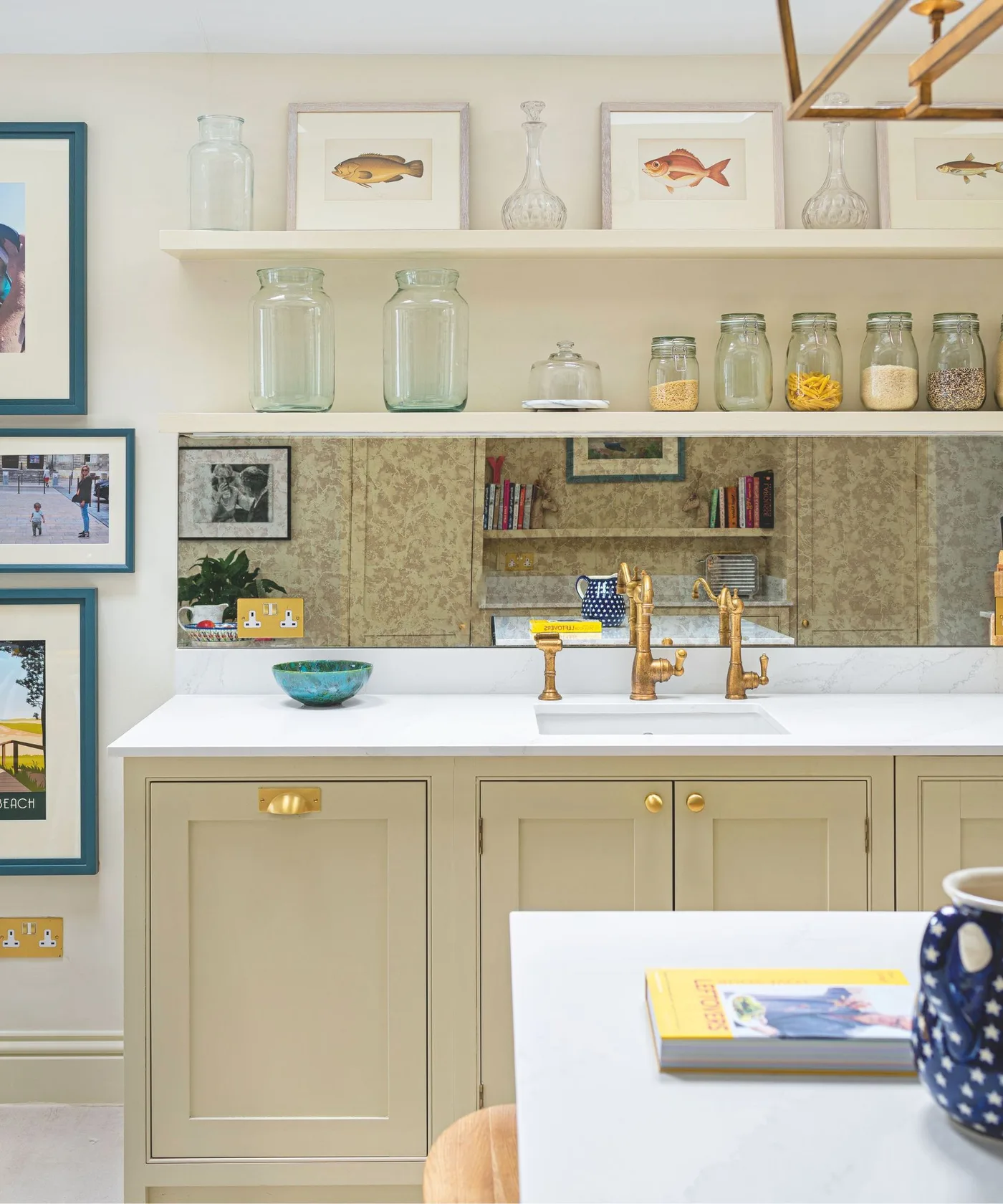 Cream kitchen cabinets with a mottled reflective backsplash, white sink, brass-colored taps and open shelves holding jars.