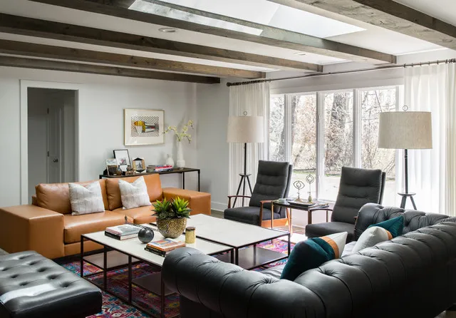 A dark tufted sofa sits opposite warmer seating beneath pale walls and timber ceiling details.