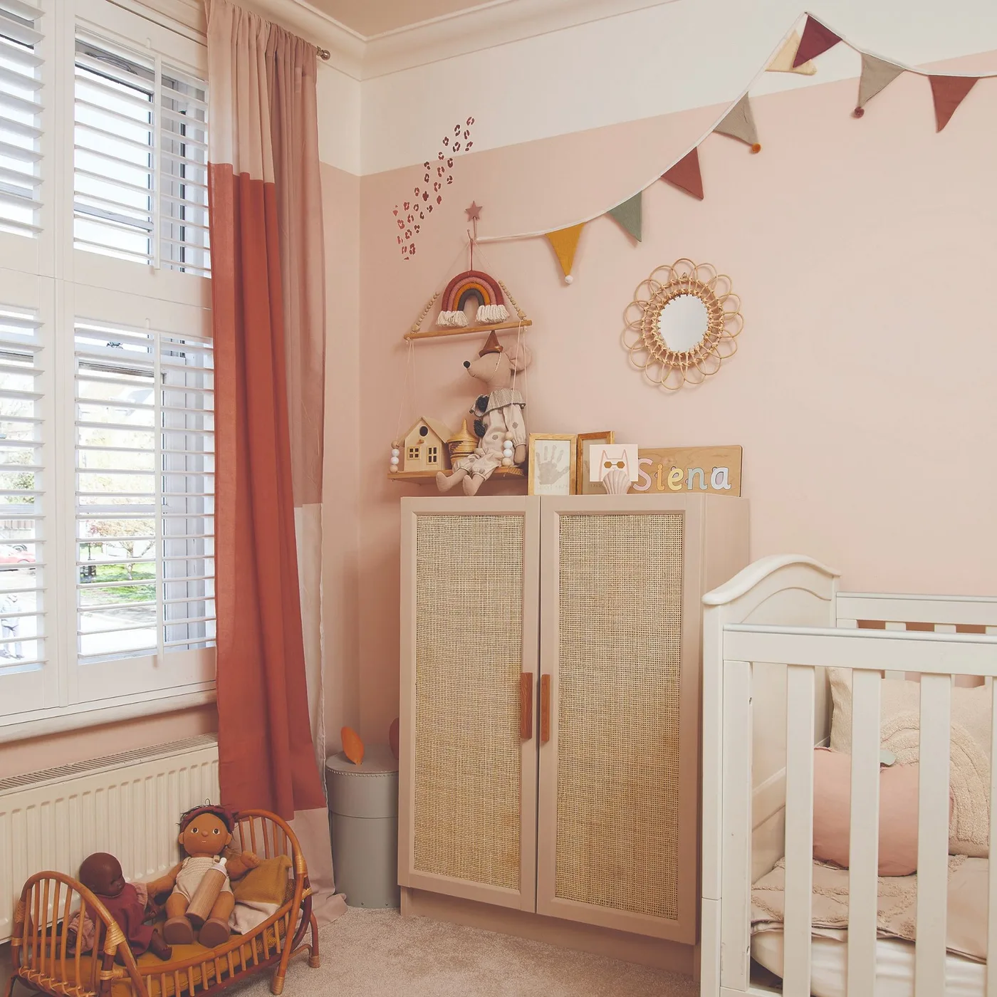Pink nursery with white crib, woven-looking cupboard fronts, decorative round mirror, smaller wall pieces and pale shutters.