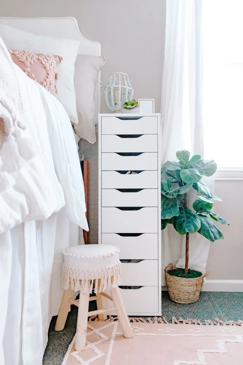 Tall narrow white drawer unit beside a raised student bed, with a small pale stool, woven plant basket and pink rug.