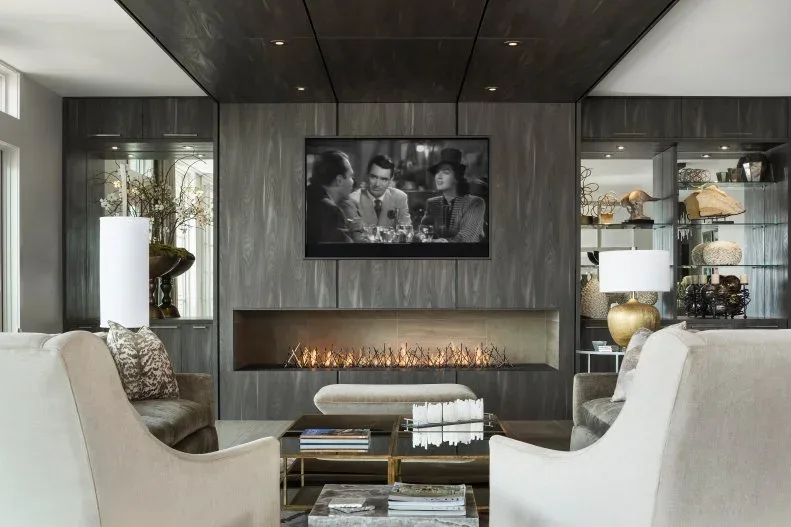 Dark gray fireplace wall with television, glass-shelved side displays and pale upholstered chairs
