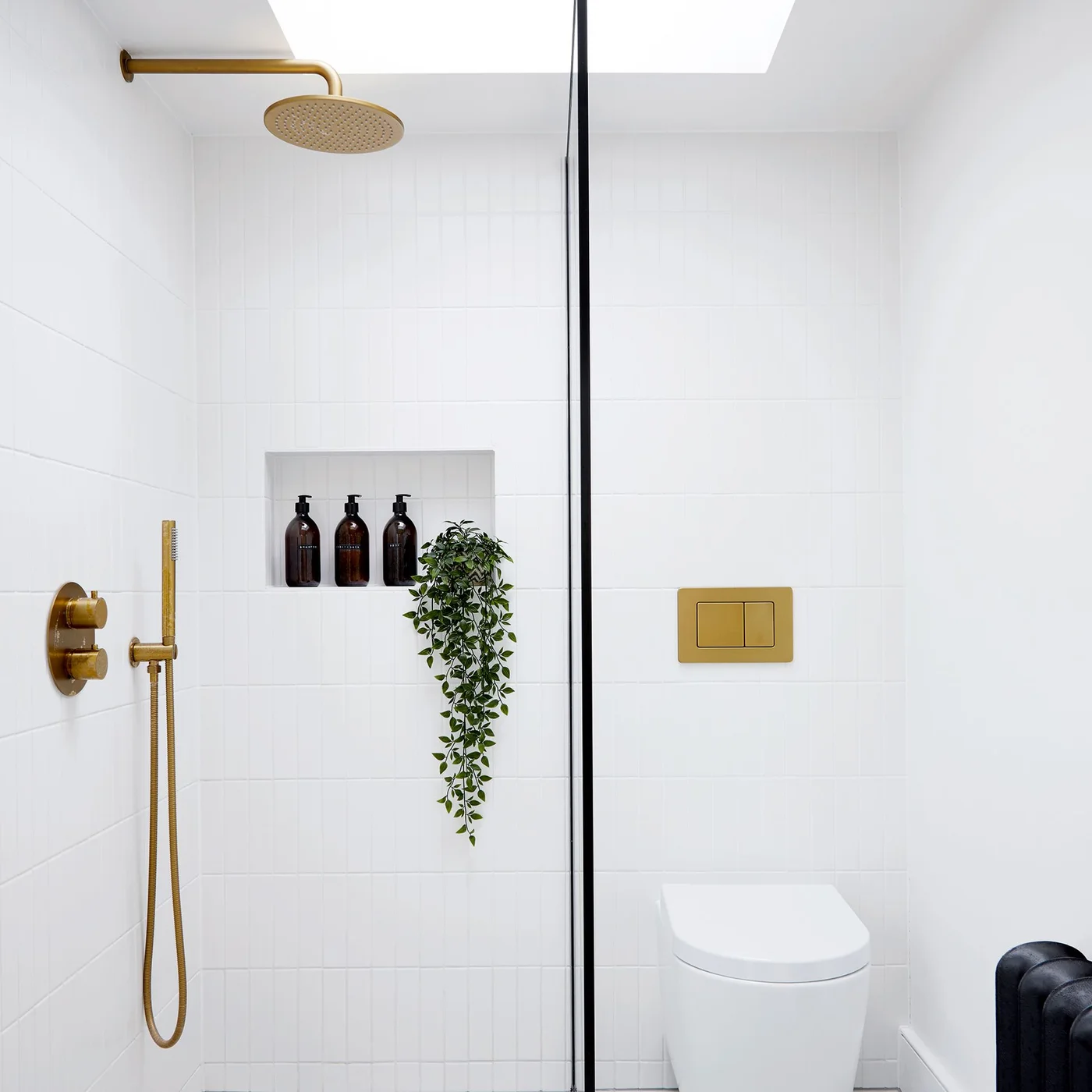 A white tiled shower has gold-colored fittings, a recessed niche and a slim dark glass-screen edge beside a white toilet below a skylight