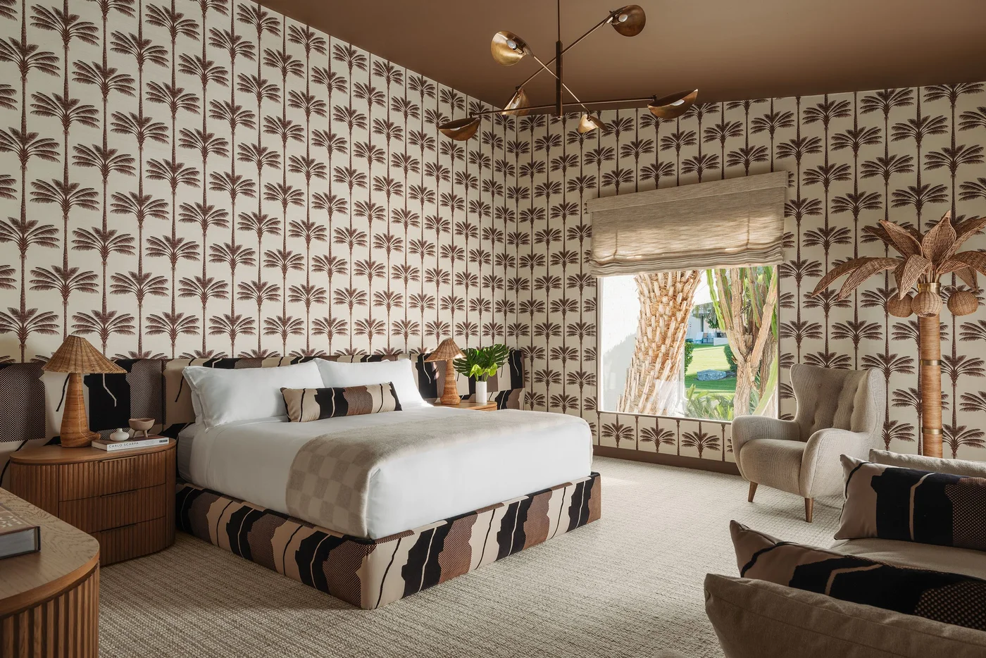 Repeated brown palm-tree wallpaper surrounds a bedroom with a patterned bed base, white bedding, rounded wood nightstands and a pale armchair