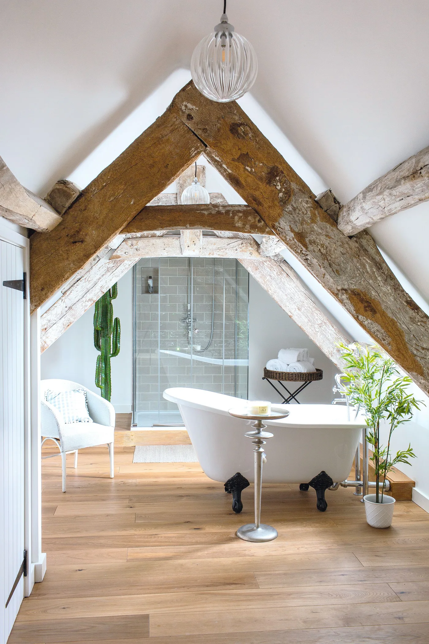Country bathroom beneath large angled beams with a white freestanding bath, separate glazed shower, pale chair and plants.