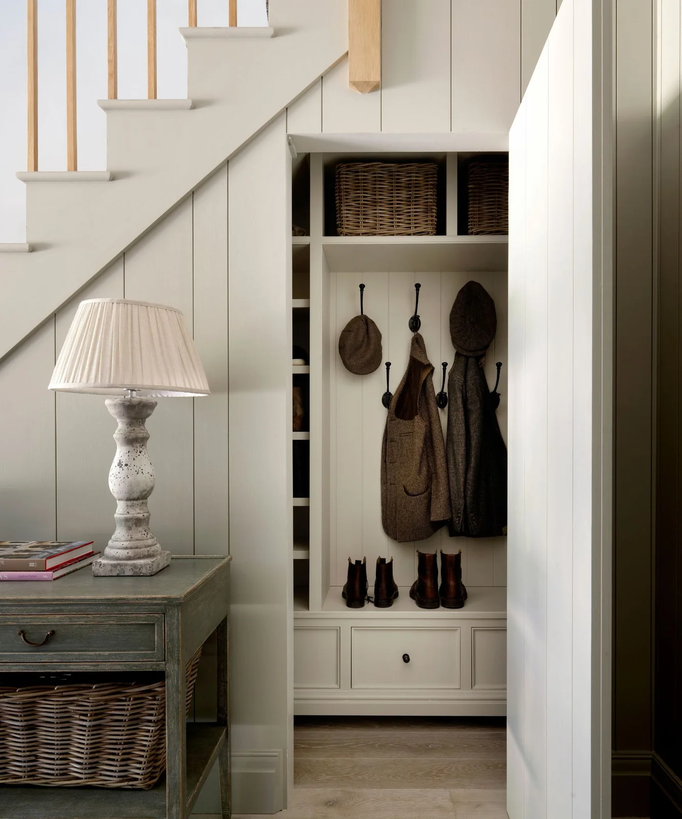 Pale stair panels conceal an open coat-storage bay with hooks, baskets and lower drawers beside a rustic side table and shaded lamp