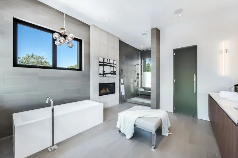 A gray bathroom with a rectangular white tub and glass shower has a small black-framed fireplace opening beside a frosted green door