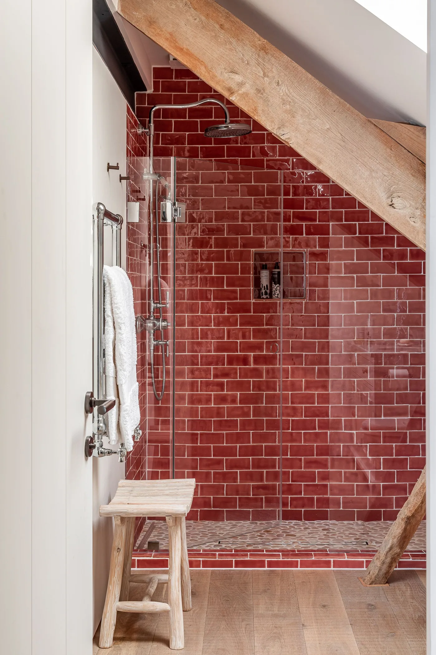 Country shower room with glossy red rectangular tiles, a glass screen, overhead beam and small tiled niche for bottles.