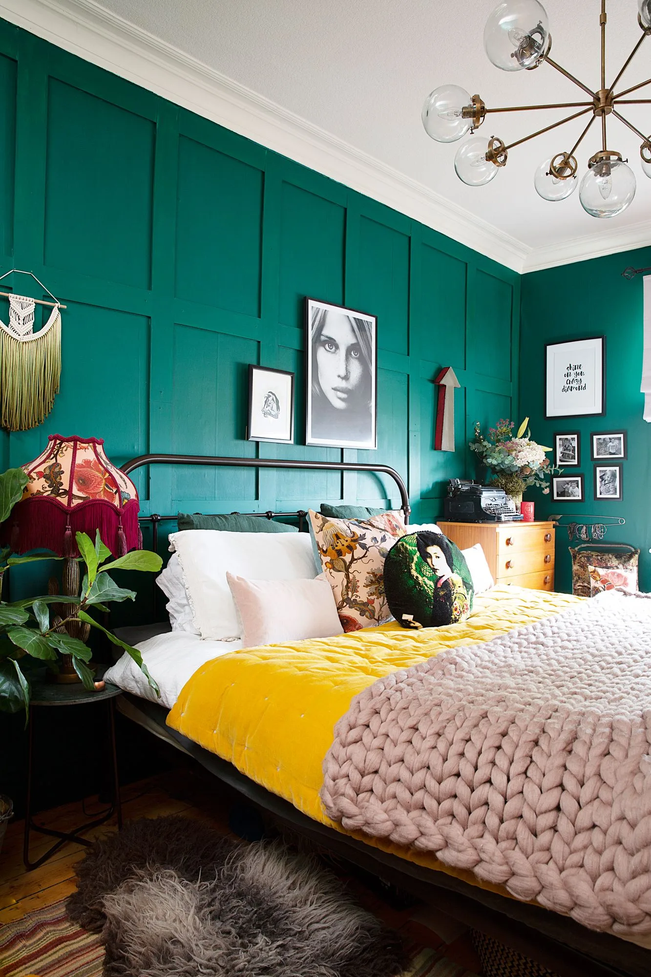 Emerald paneled bedroom with a dark metal bed, white pillows, yellow throw, pink knitted blanket, portraits, and plants.