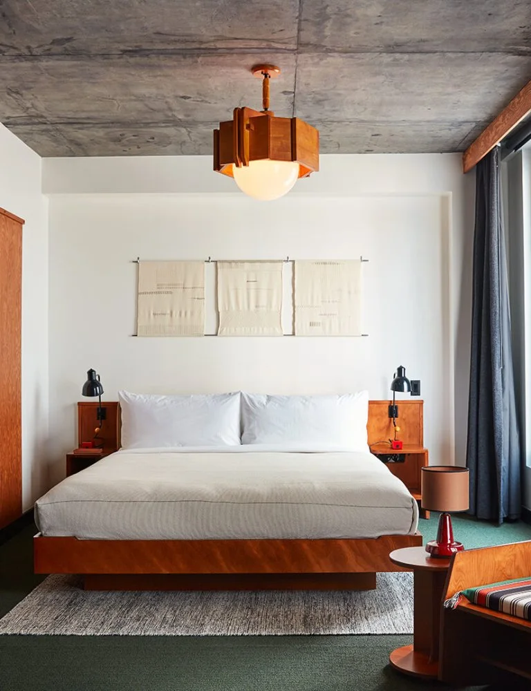 A wood platform bed stands beneath a wood-framed geometric pendant beside warm bedside units, pale textiles, dark curtains and green bedroom carpet