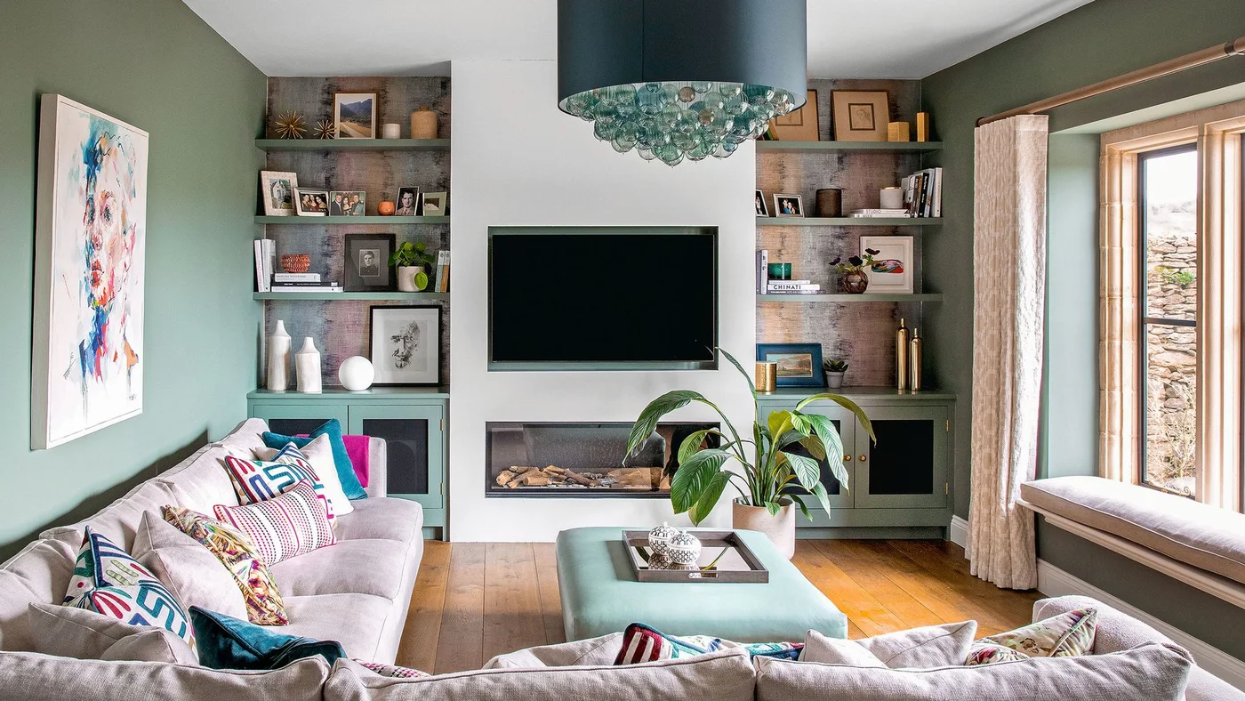 A television above a long fireplace is framed by open shelves and green lower cabinets beside pale sofas and a window seat