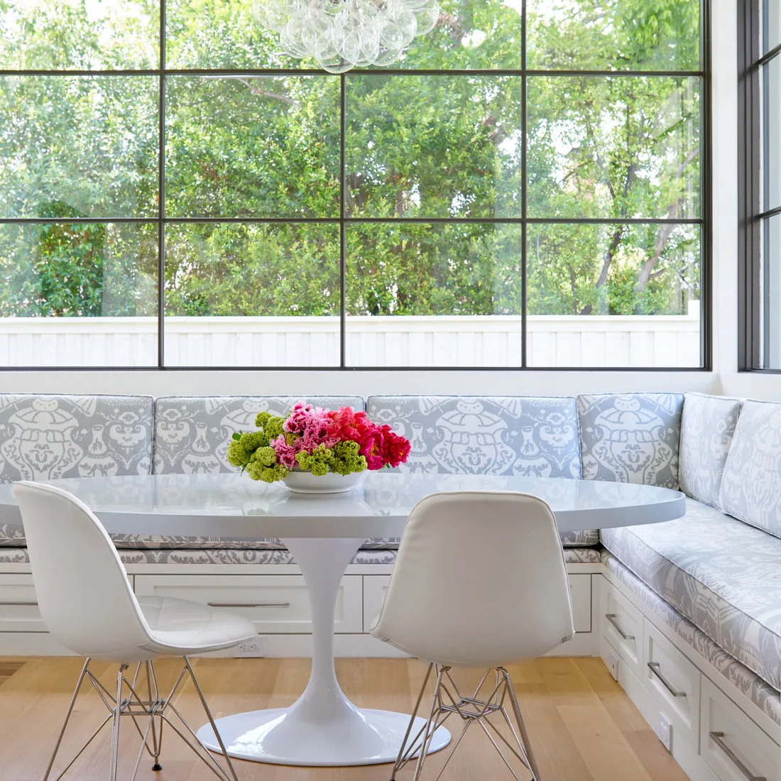 White oval pedestal table with white chairs and a pale patterned corner banquette below large windows