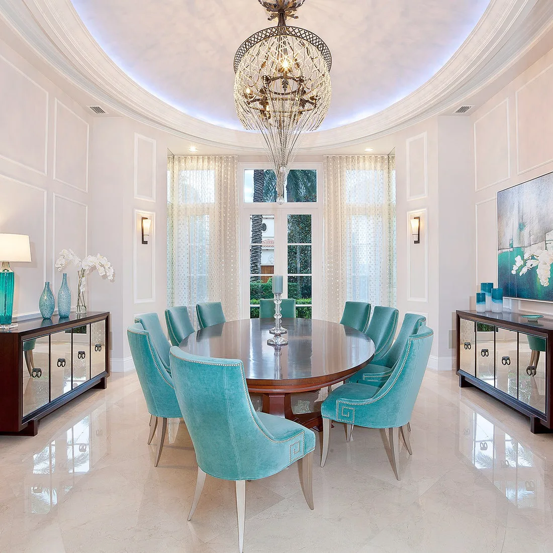 Dark oval dining table with turquoise upholstered chairs between two sideboards beneath an ornate chandelier