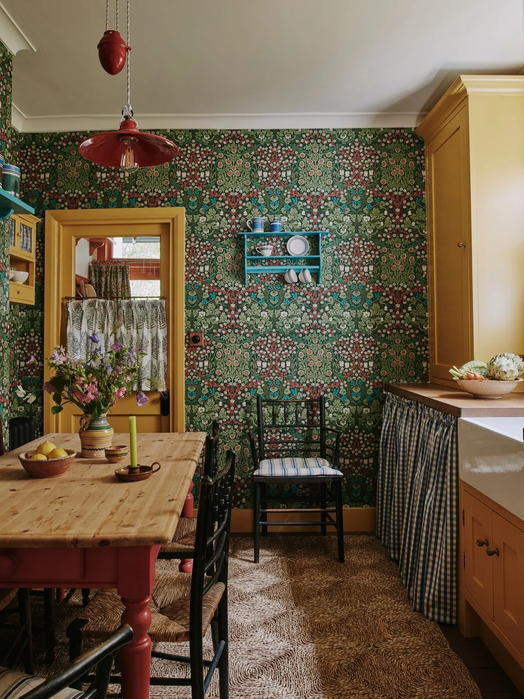 Dense green floral wallpaper surrounds yellow trim, a red pendant, turquoise wall shelf and checked fabric beneath a kitchen counter beside a wooden table