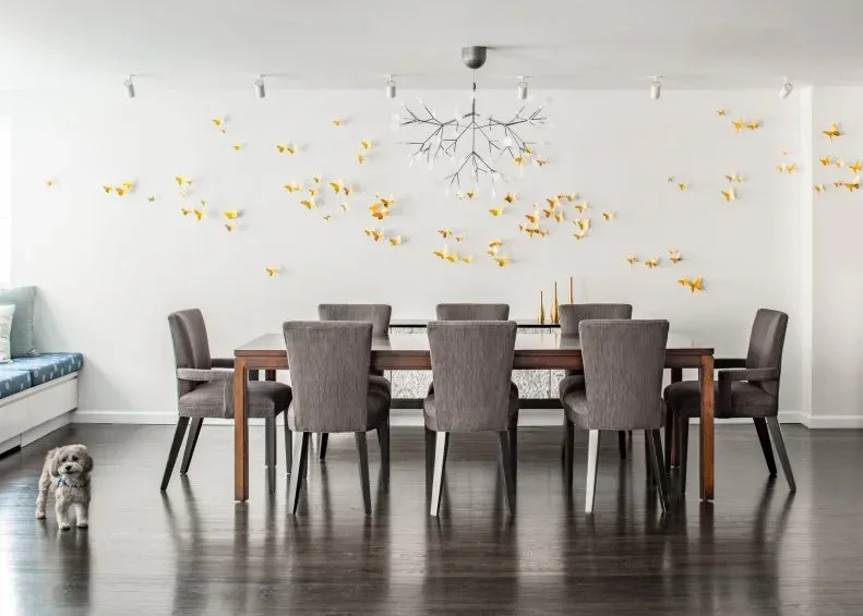 Slender dark branch-like chandelier above a long table and gray chairs, against a pale wall decorated with scattered yellow butterflies and ceiling track lights.