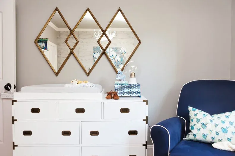 A white nursery dresser with dark handles stands beneath three diamond-shaped mirrors beside a blue upholstered chair and a small changing surface