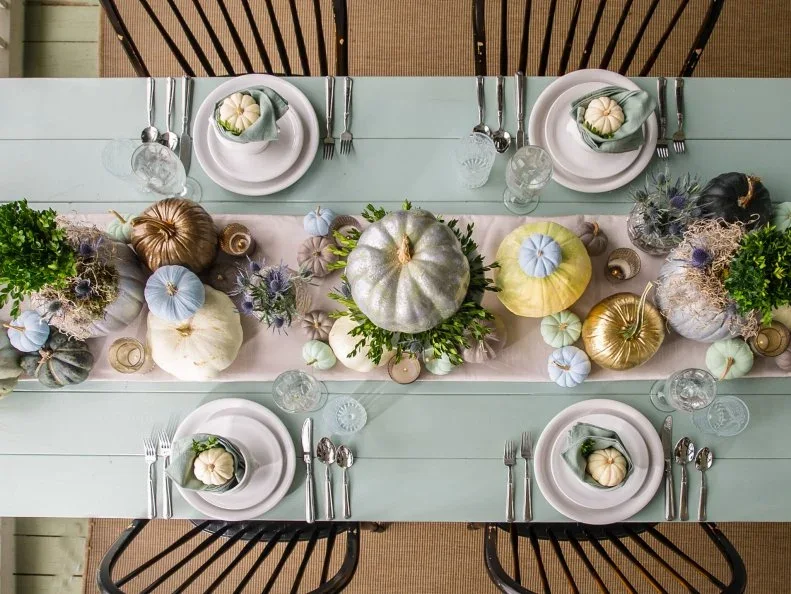 A sage-green tabletop holds pale plates, green napkins and small pumpkins around a pale runner with painted and metallic pumpkins