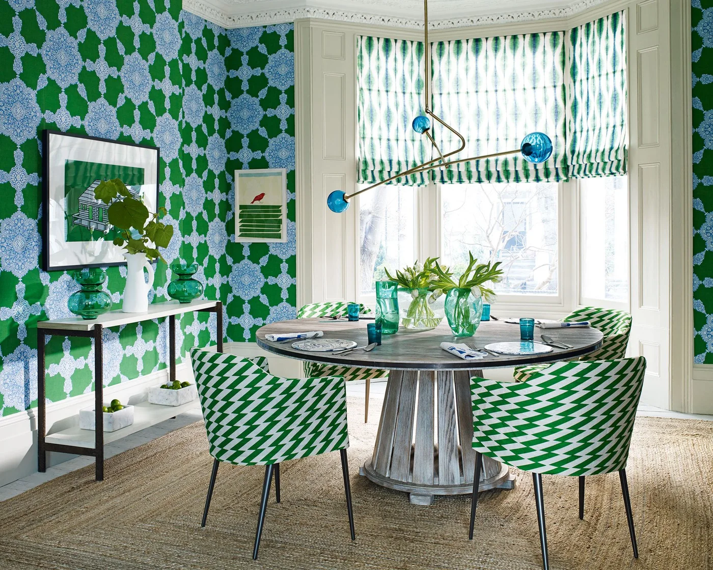 Green-and-pale patterned chairs surround a dark round table beneath blue and green wall patterns and a sculptural blue pendant