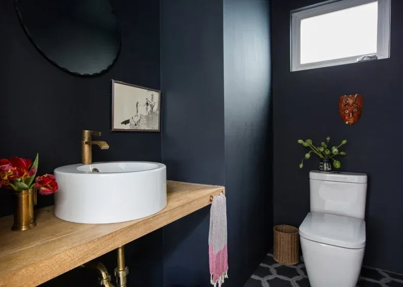 Powder room with deep blue walls, white bowl basin on wooden counter, dark round mirror, framed drawing, small red wall object and white toilet.