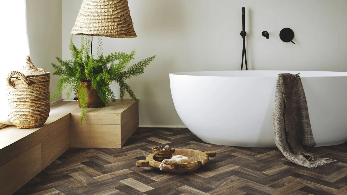 A white oval bath stands above a brown herringbone wood-look floor beside a woven shade, pale walls and black wall-mounted fittings