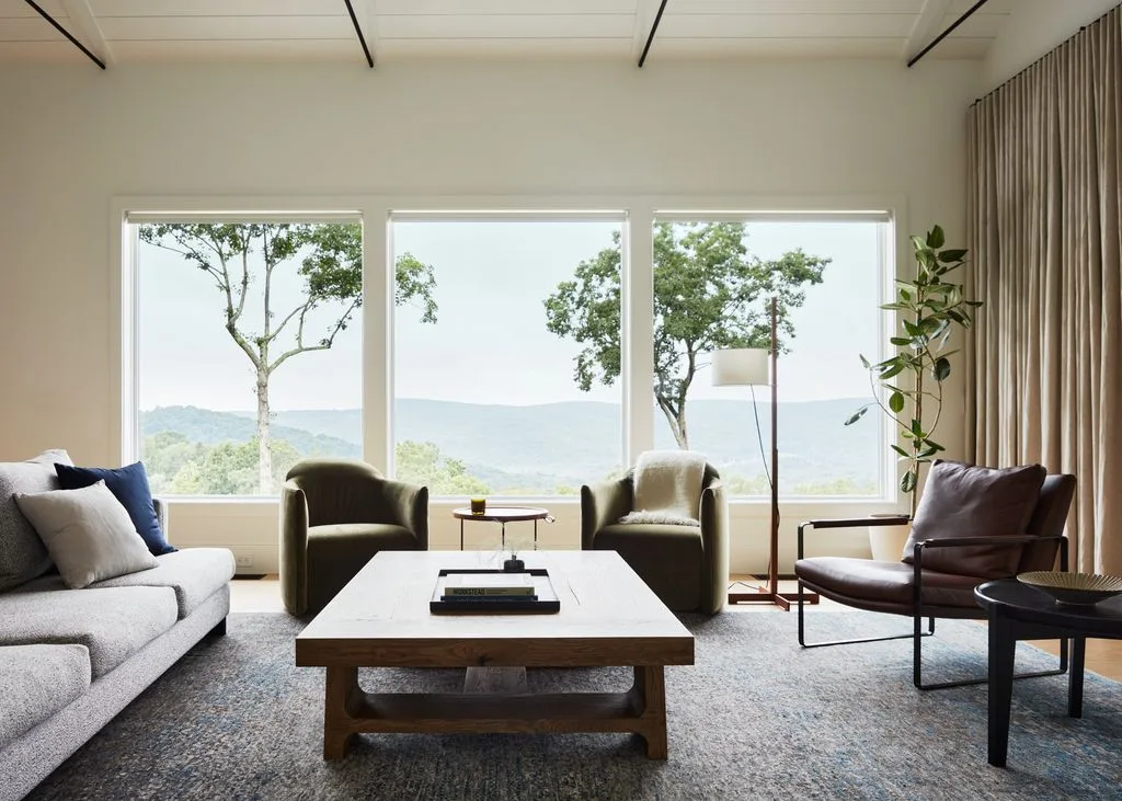 Living room with a pale sofa, two muted green armchairs, wooden coffee table, long curtains and a broad landscape-facing window.