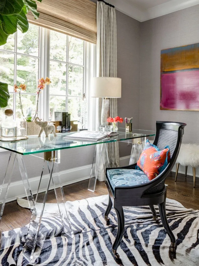 Glass-topped desk on clear supports beside a black curved chair, patterned rug and colorful artwork.