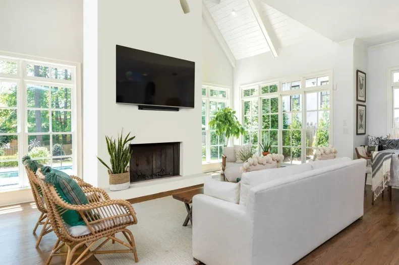 A white sofa and two woven chairs face a pale fireplace wall and television beside broad windows above a pale rug and wood floor