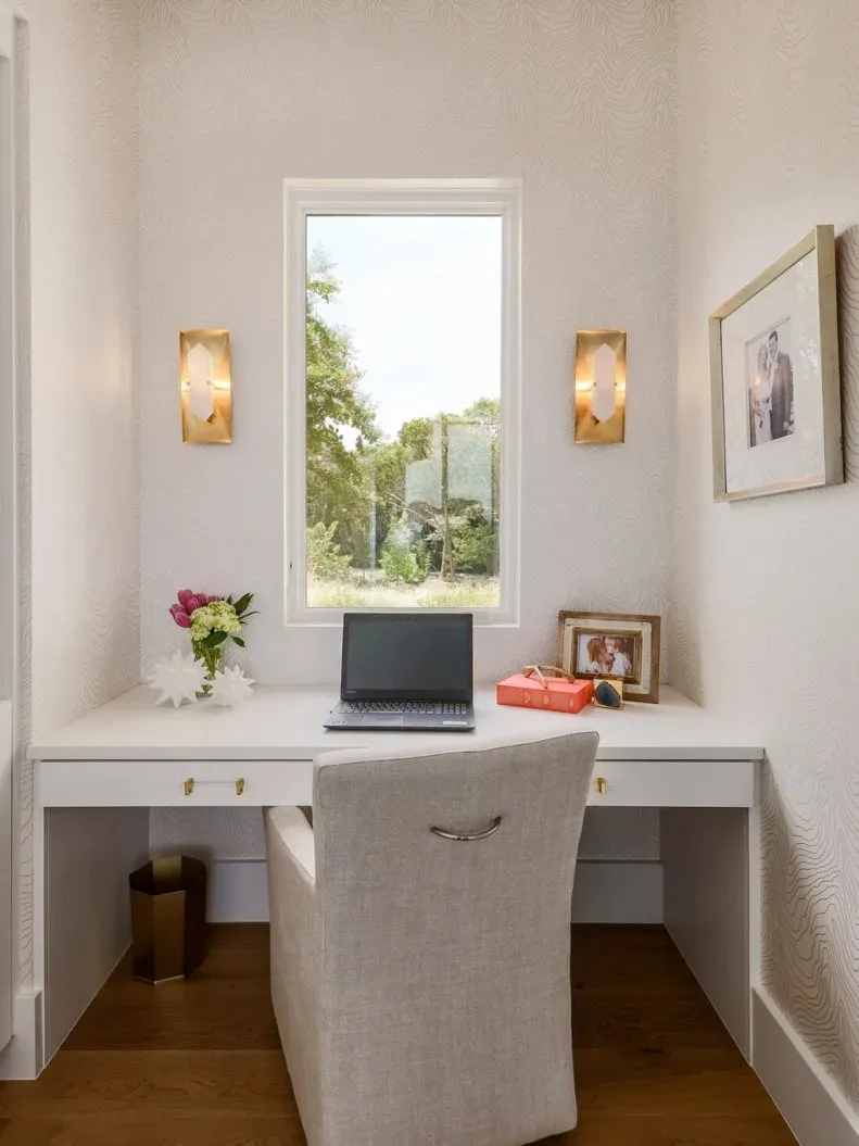 Pale built-in desk below a narrow window with two gold-toned wall sconces and a tall upholstered chair.