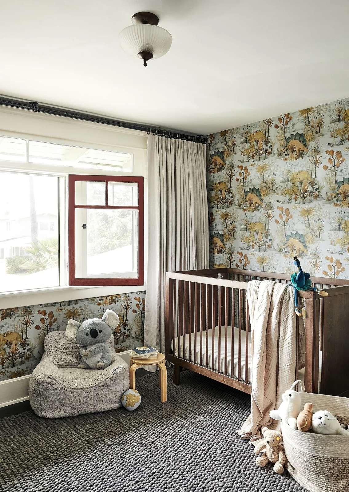 Illustrated dinosaur and plant wallpaper wraps around a nursery with a dark wooden crib, gray rounded chair, pale curtains and red window frame