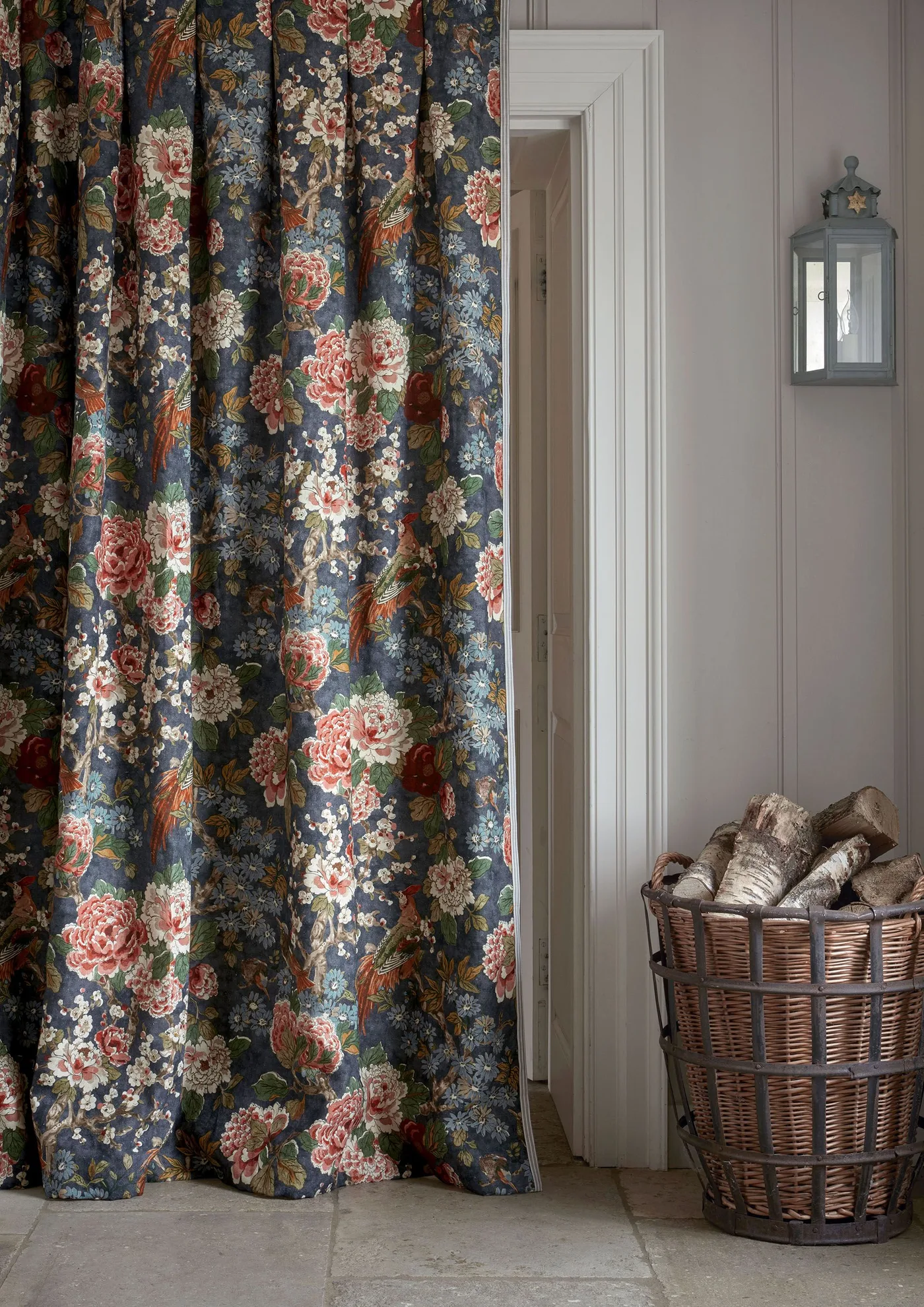 Country entrance detail with a dark floral curtain, pale gray vertical wall paneling, a wall lantern and a woven basket holding logs.