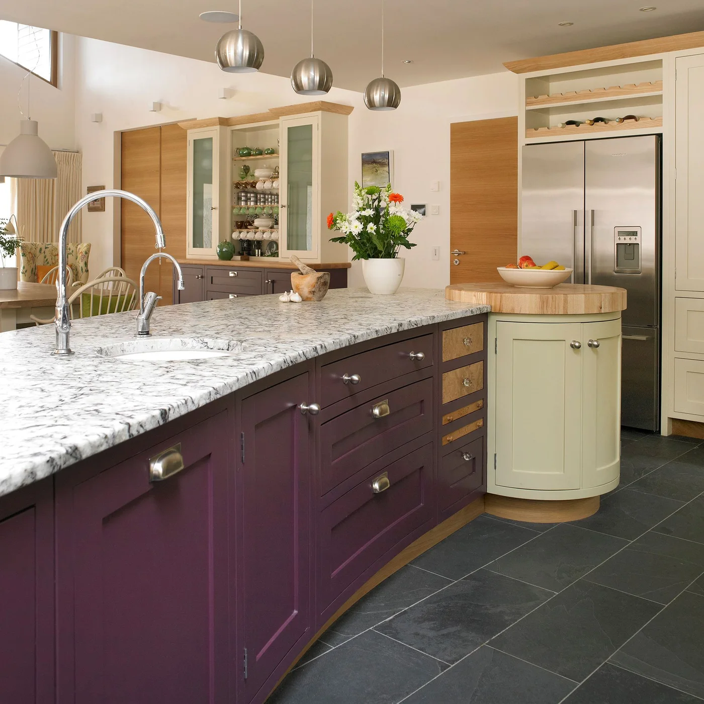 A plum-colored island with pale speckled counter and sink sits beside cream storage, open shelves and a tall refrigerator under three hanging lights