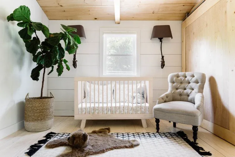 White crib and buttoned chair beneath pale wooden ceiling beside wooden wall, patterned rug and woven plant container.
