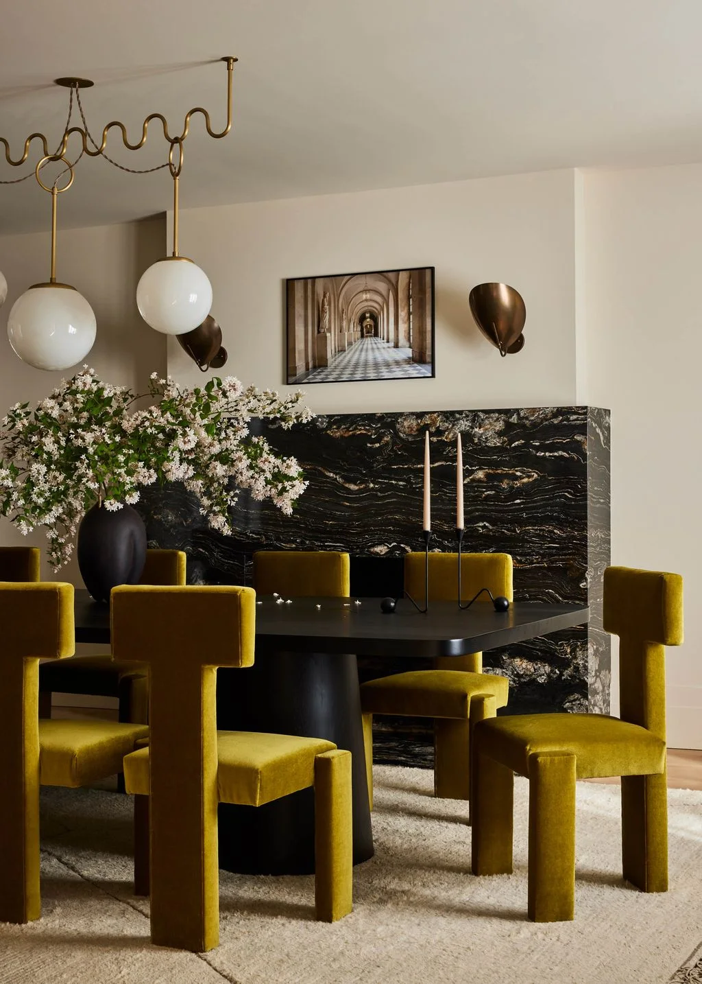 Yellow upholstered chairs surround a black dining table beneath white globe pendants beside a dark veined wall and warm metal wall lights