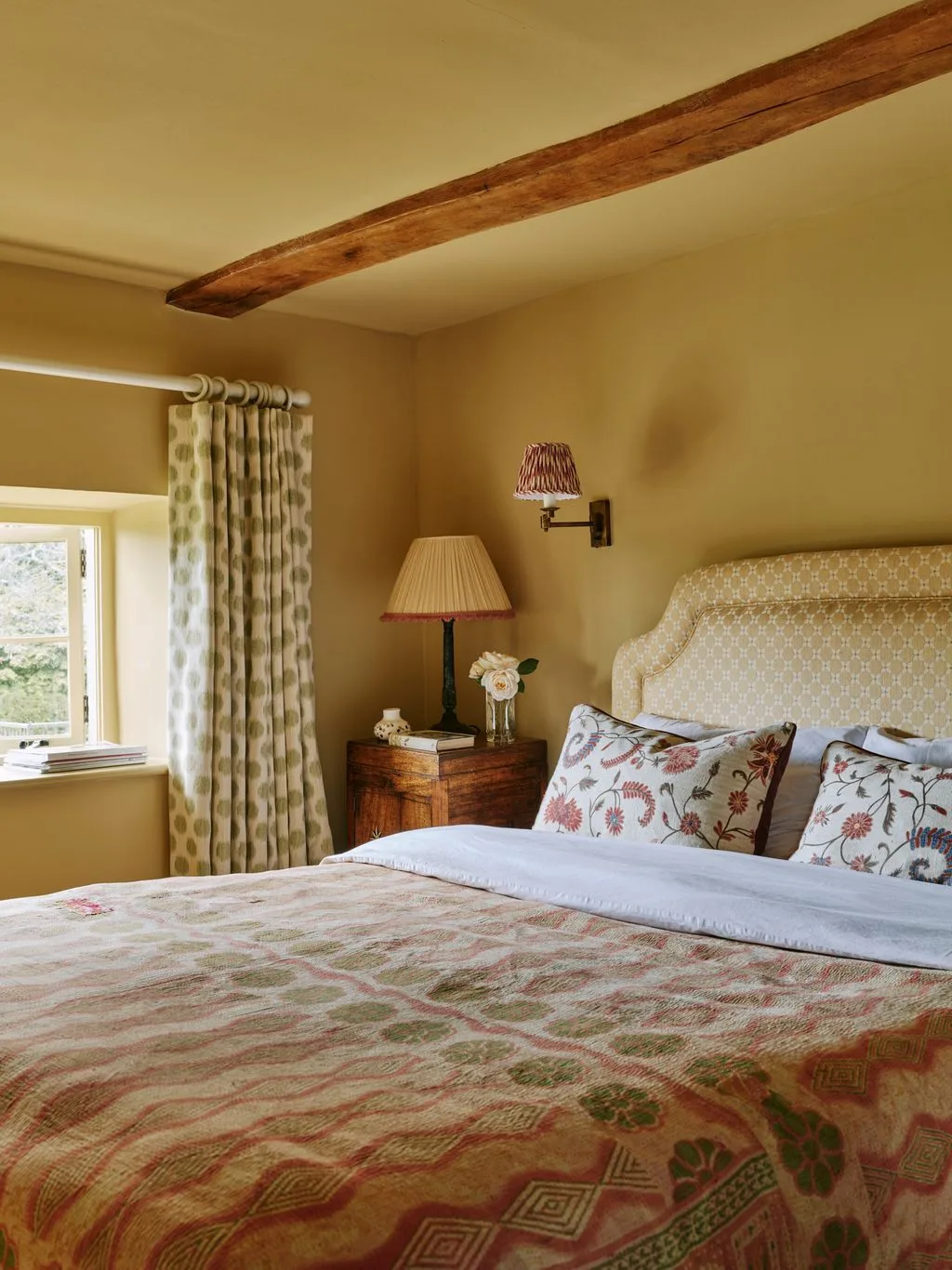 Bedroom with warm yellow walls, exposed ceiling beam, shaped pale headboard, floral cushions, patterned bed cover and checked curtains