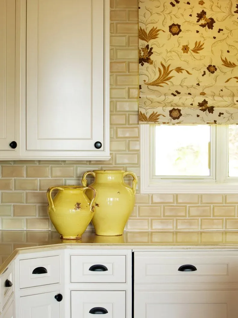 White kitchen cabinets with black hardware, pale yellow beveled rectangular backsplash tiles, yellow pitchers and a yellow floral window blind.