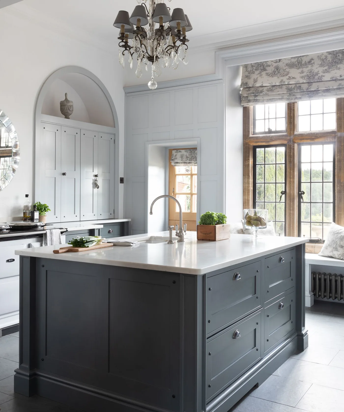 Pale kitchen with a dark paneled island, sink, tall arched cupboard and a shaded chandelier beside a large window.