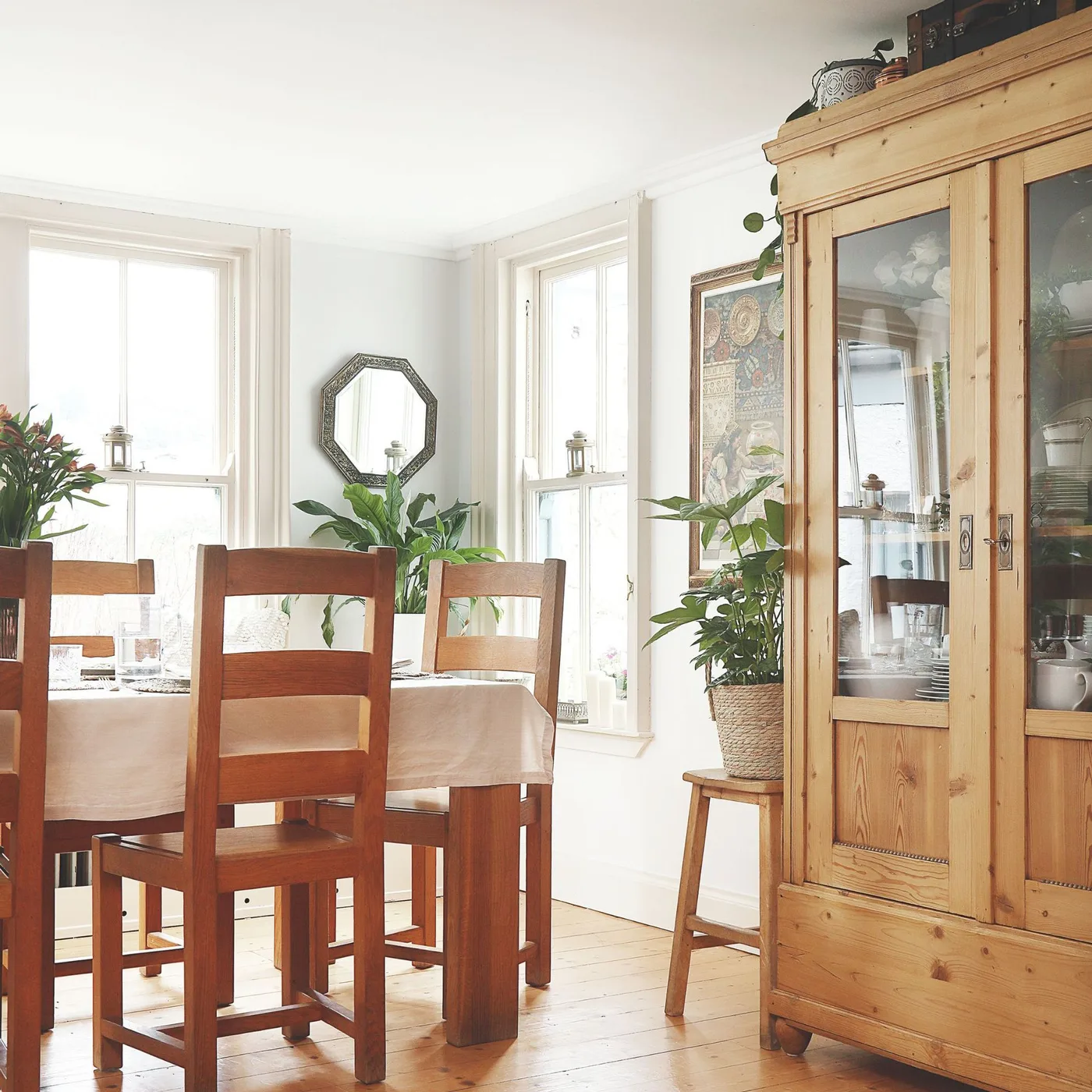 Dining room with a wooden table, wooden chairs, white tablecloth, tall glazed dresser, plants and two windows.