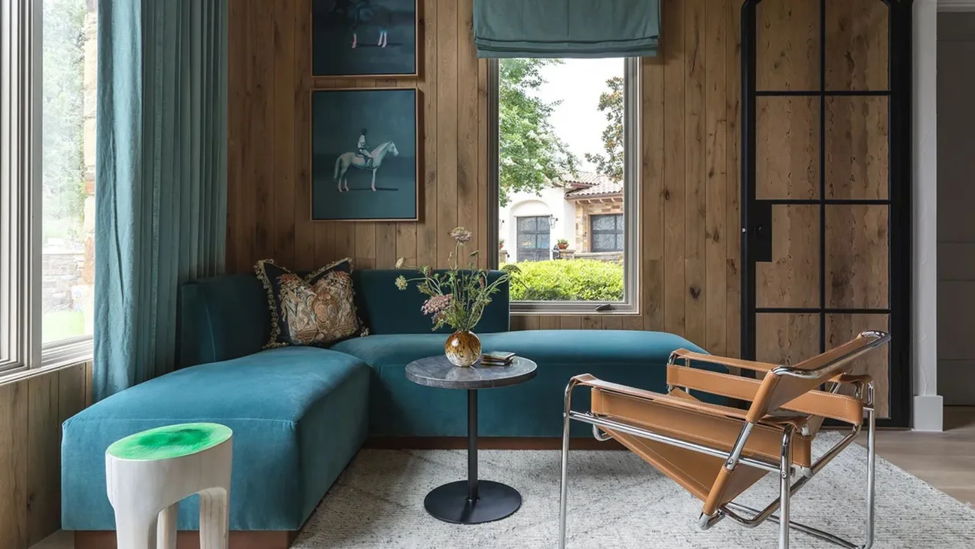 A blue corner sofa sits beside wood wall panels, a small round table and an open-framed brown chair near tall windows