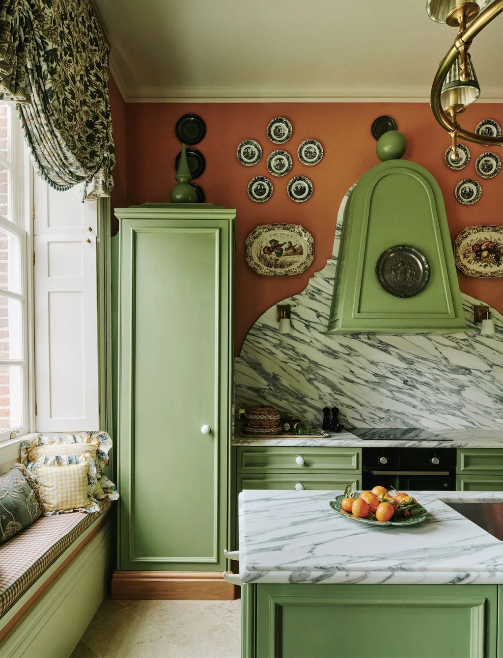 Green cabinets and a curved hood surround a strongly veined backsplash beneath decorative plates on an orange wall beside patterned curtains