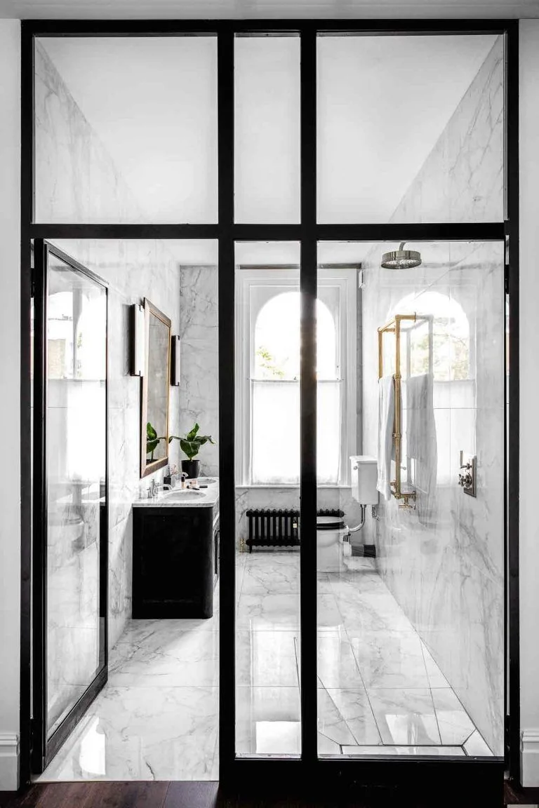 Black-framed glazing opens toward a white veined bathroom with a dark vanity, black radiator and white freestanding tub