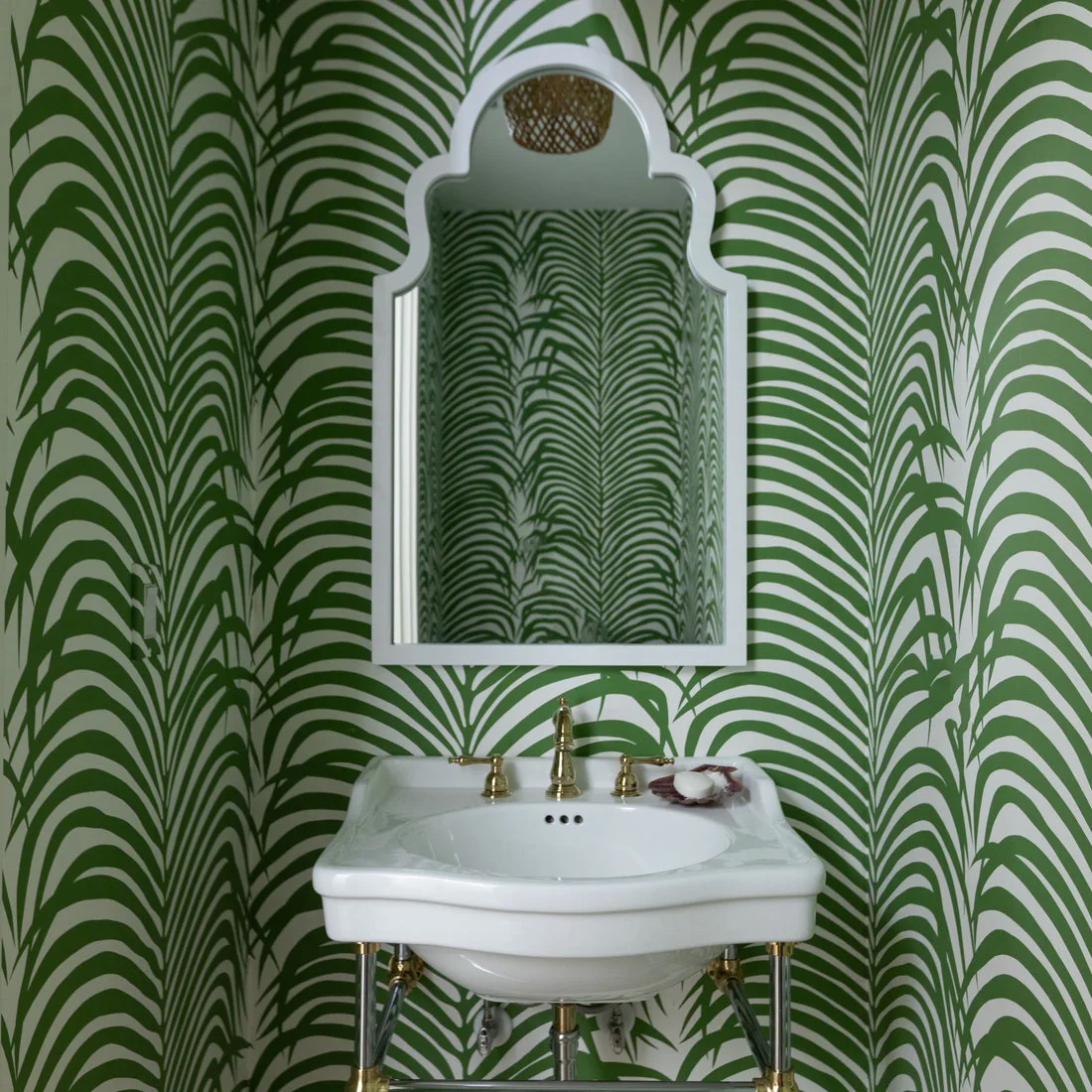 Large green-and-white palm patterns surround a shaped white powder-room mirror above a pale console sink and gold-colored fittings