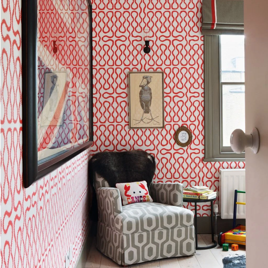 Red looping nursery wallpaper surrounds a gray geometric adult chair beside framed bird pictures, a dark side table and window blind