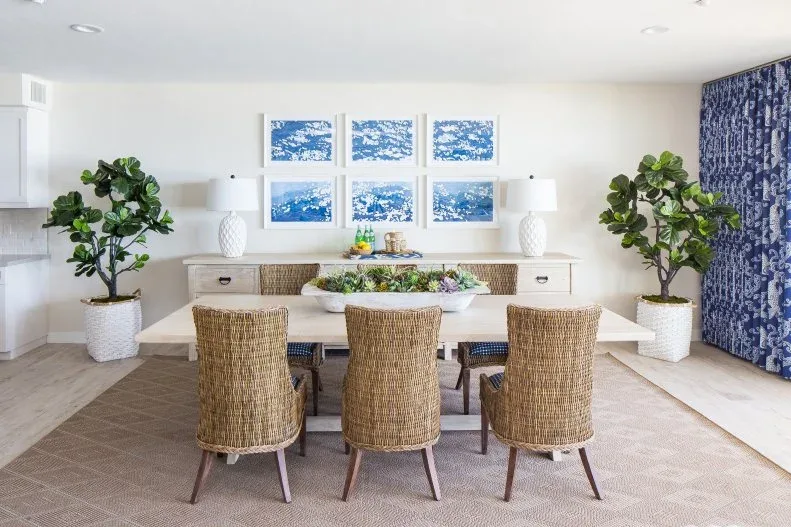 Tall woven-back dining chairs around a white table, with blue-patterned curtains, blue framed pictures, pale cabinetry, and large plants.