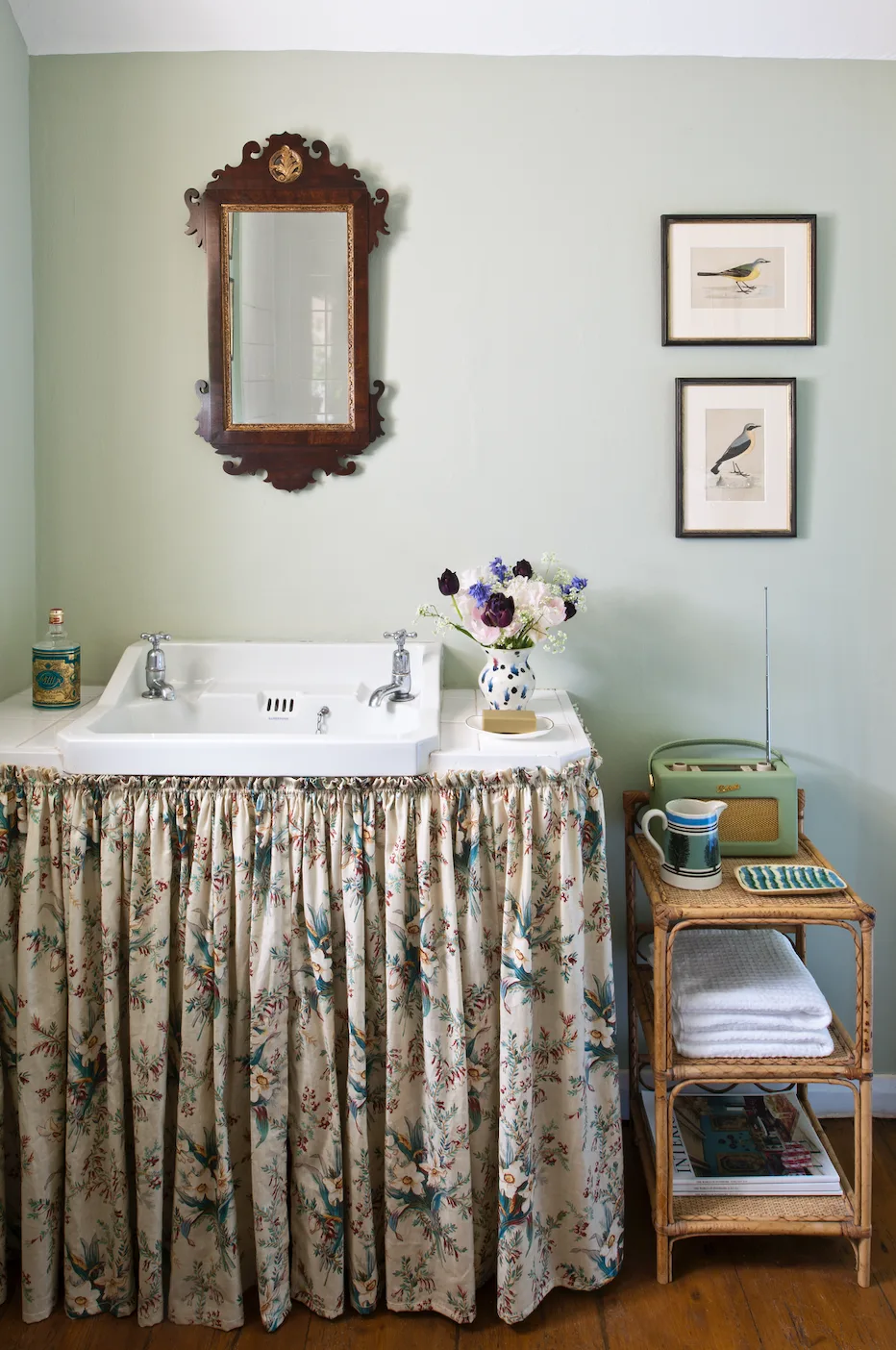 Country bathroom with a floral skirt beneath a white basin, pale green walls, ornate wooden mirror, bird prints and a small woven shelf.