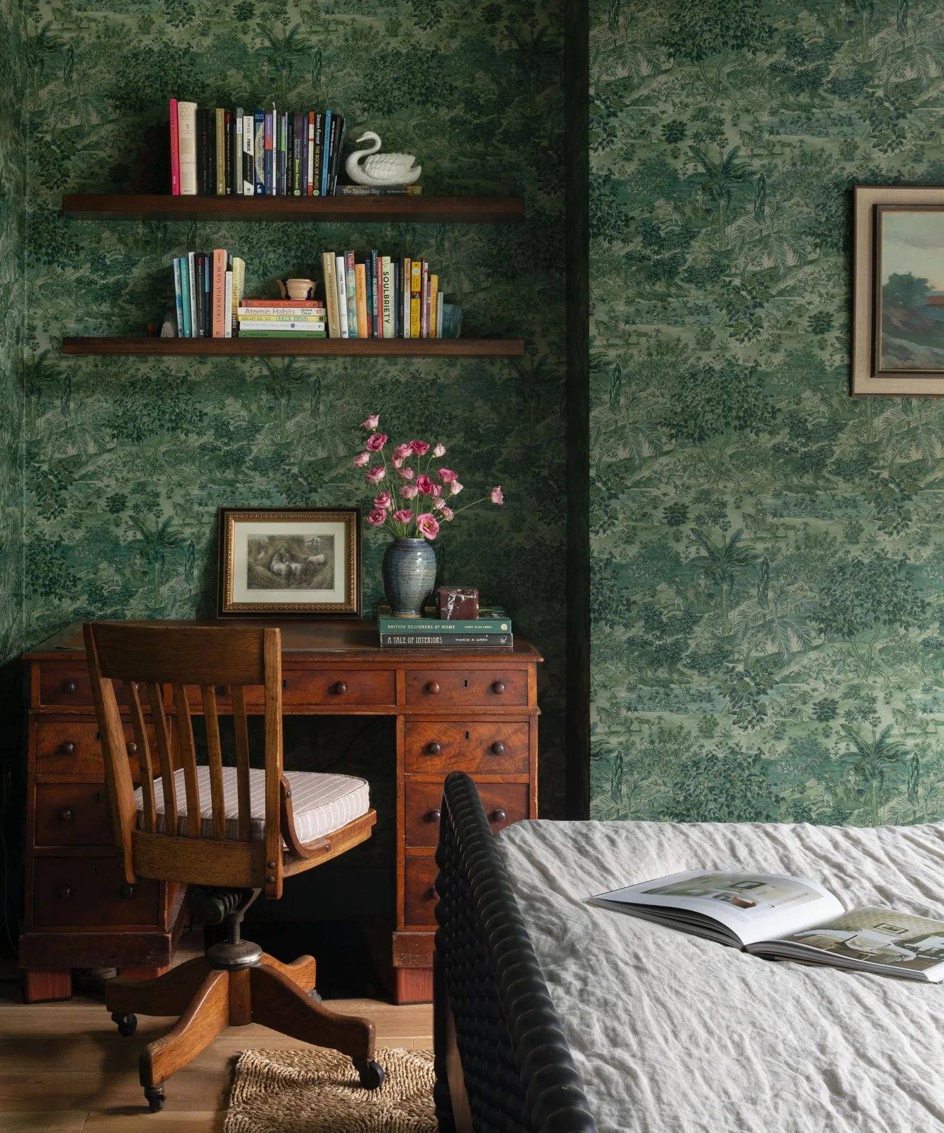 A wooden drawer desk and wooden chair sit against green patterned wallpaper beside a bed beneath open book shelves