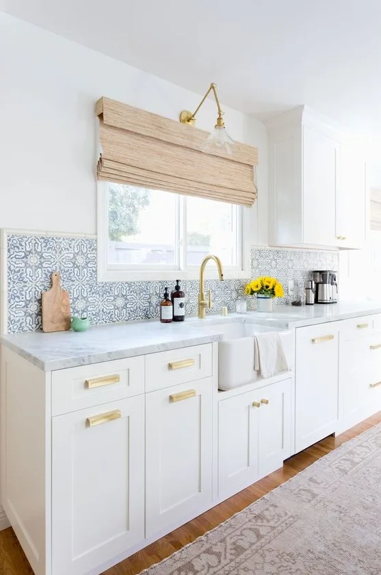 White cabinets and an apron-front sink sit beneath a blue-and-white patterned backsplash beside a warm-colored faucet, wall light and window blind