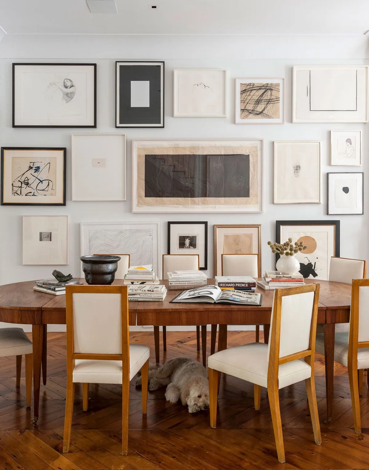 Assorted pale and dark framed artworks cover a dining-room wall behind a wooden table and pale upholstered chairs with warm-colored frames
