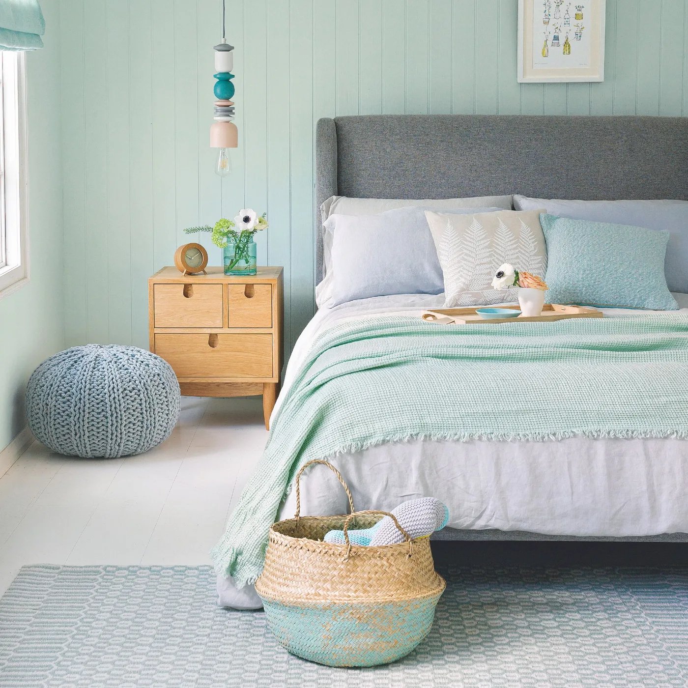 Aqua-paneled bedroom with a gray bed, pale bedding, wooden bedside cabinet, knitted footstool and woven basket.