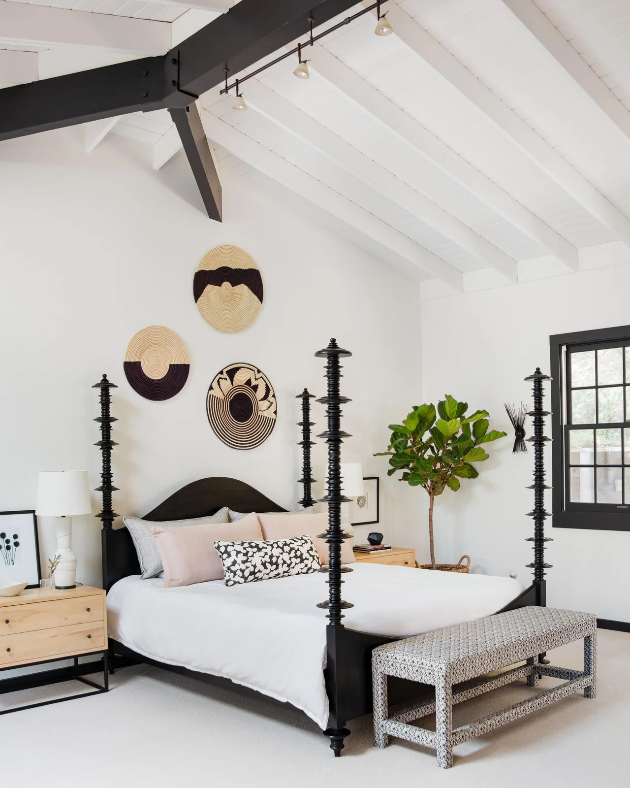 A white bedroom has a dark tall-post bed with white bedding, pale wood bedside drawers, a patterned bench and small pink cushions